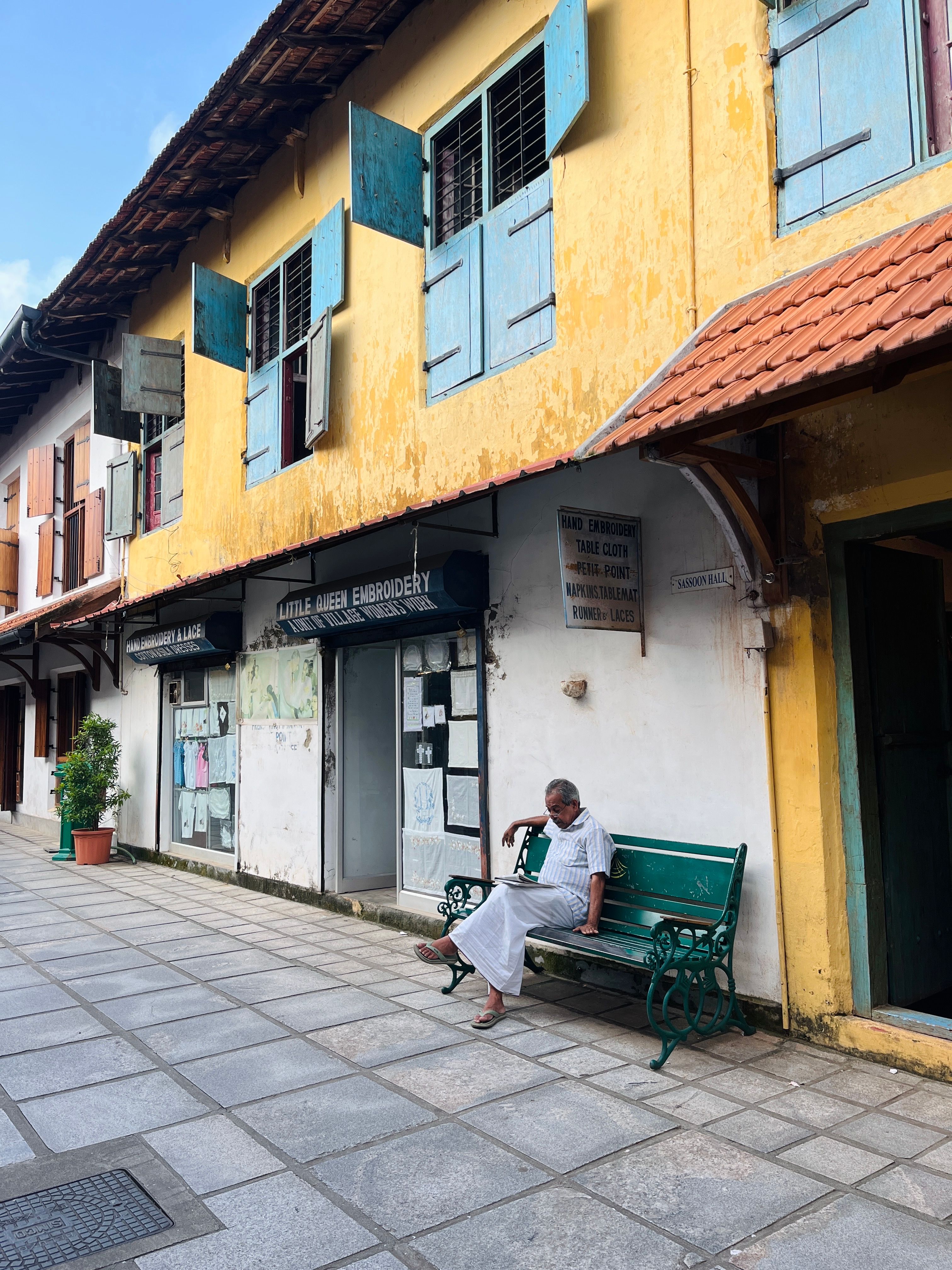 A person sits on a green bench outside a yellow-and-white building in Cochin.