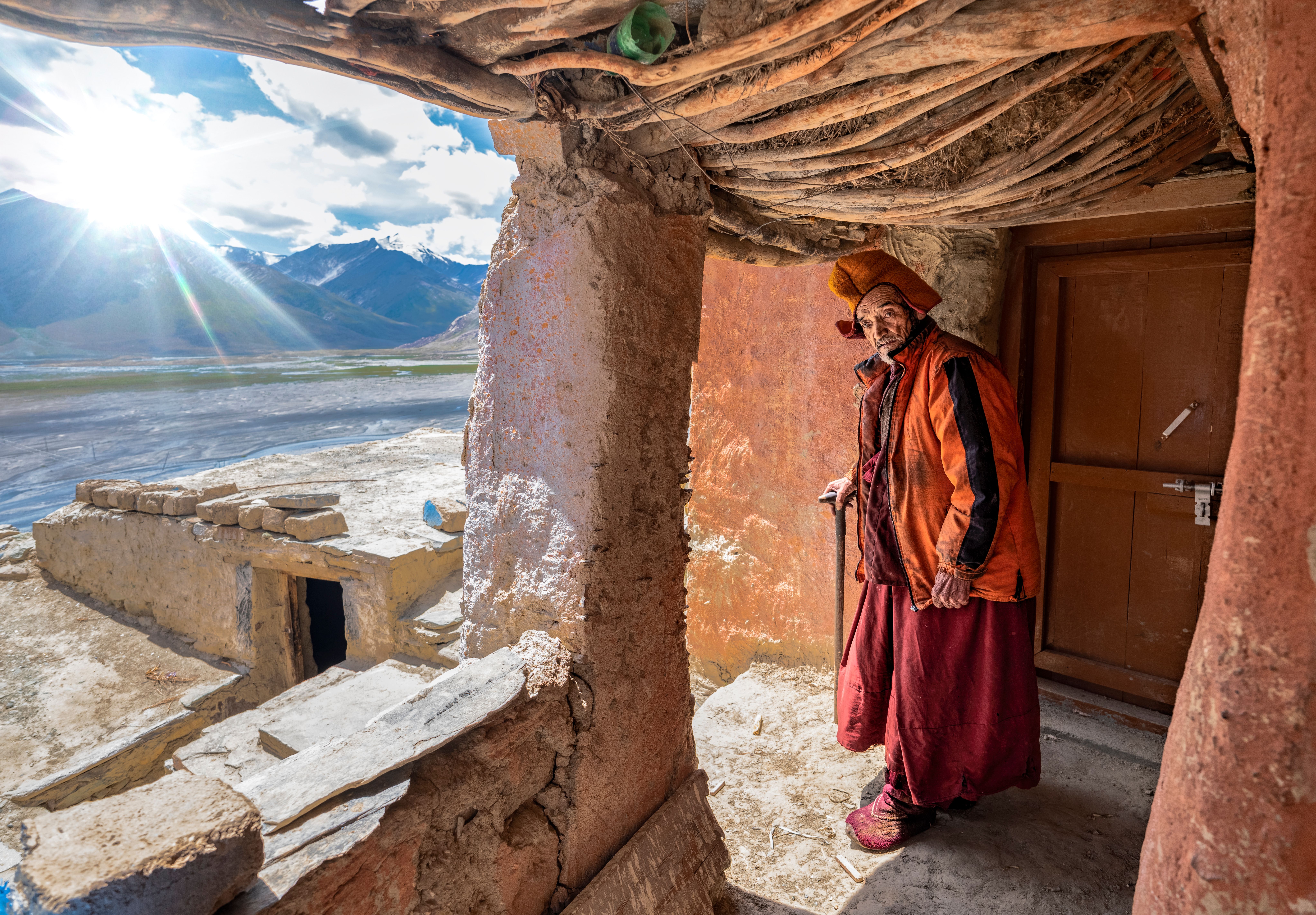 A Ladakhi elder in traditional dress stands beneath a rustic shelter overlooking a mountain valley