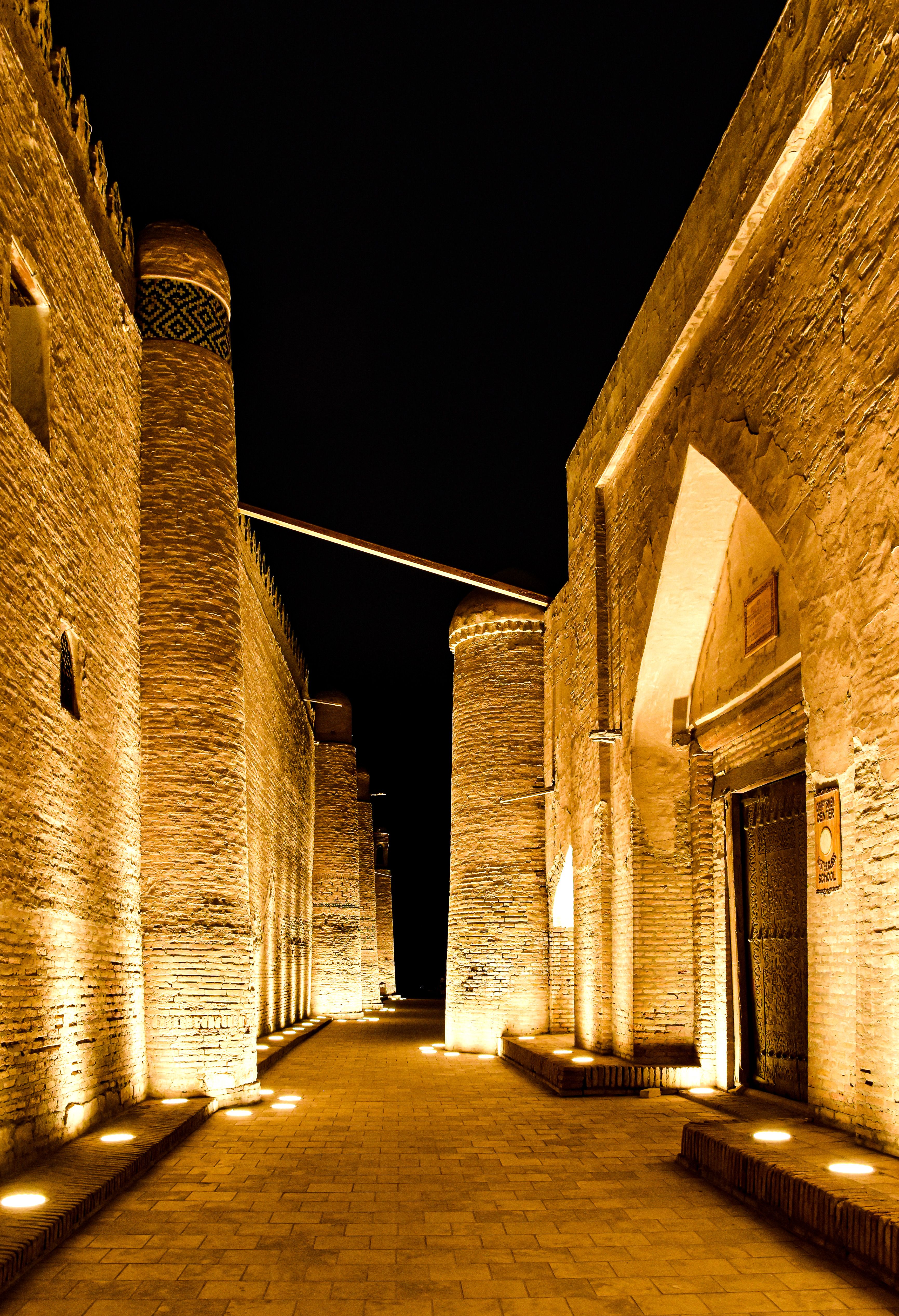 A lantern-lit lane between Khiva’s historic brick buildings, with rounded towers rising above the walls.