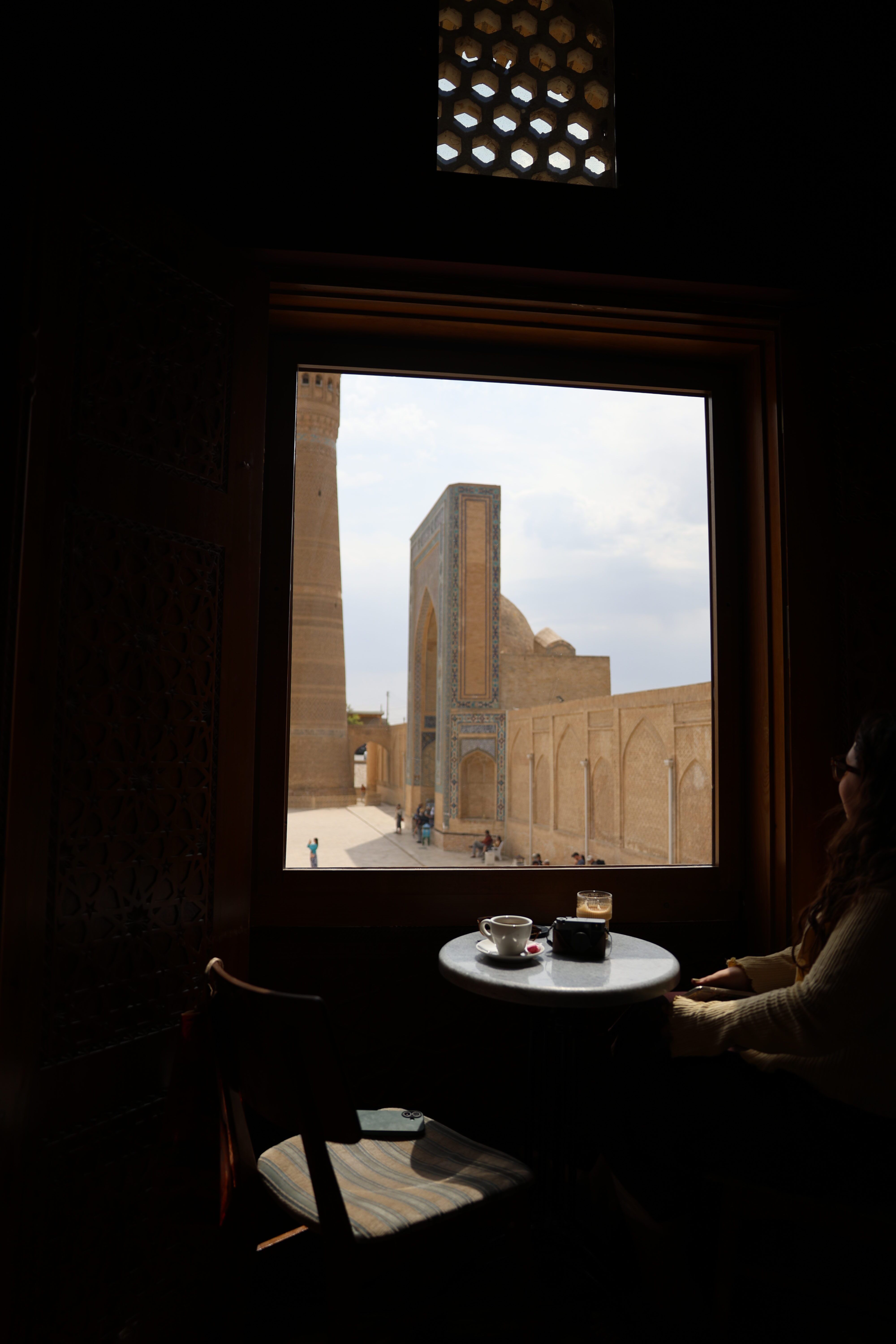 A tall brick minaret and historic buildings in Bukhara, viewed from a café window.