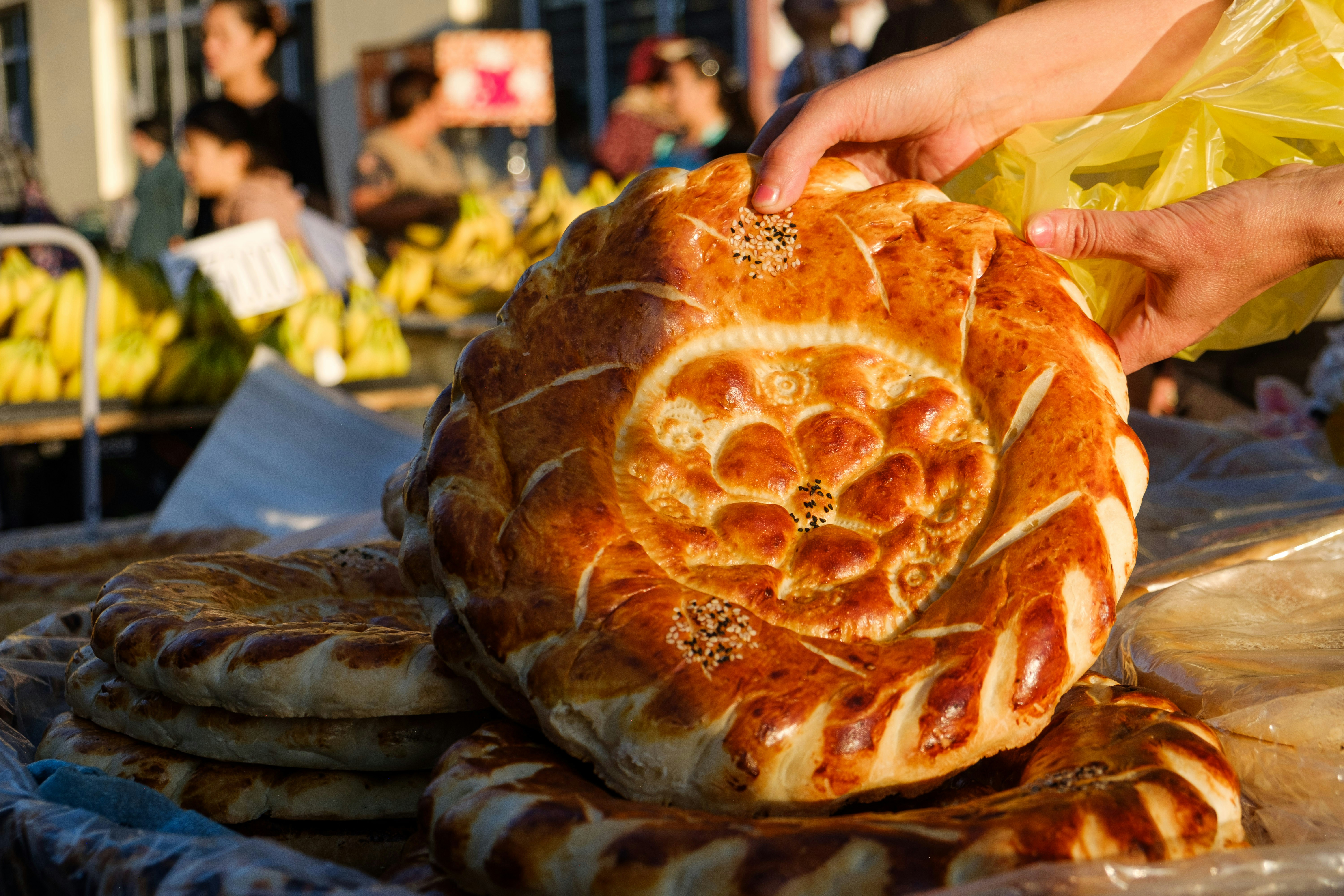 Hands selecting a golden round loaf of Uzbek bread at a market stall.