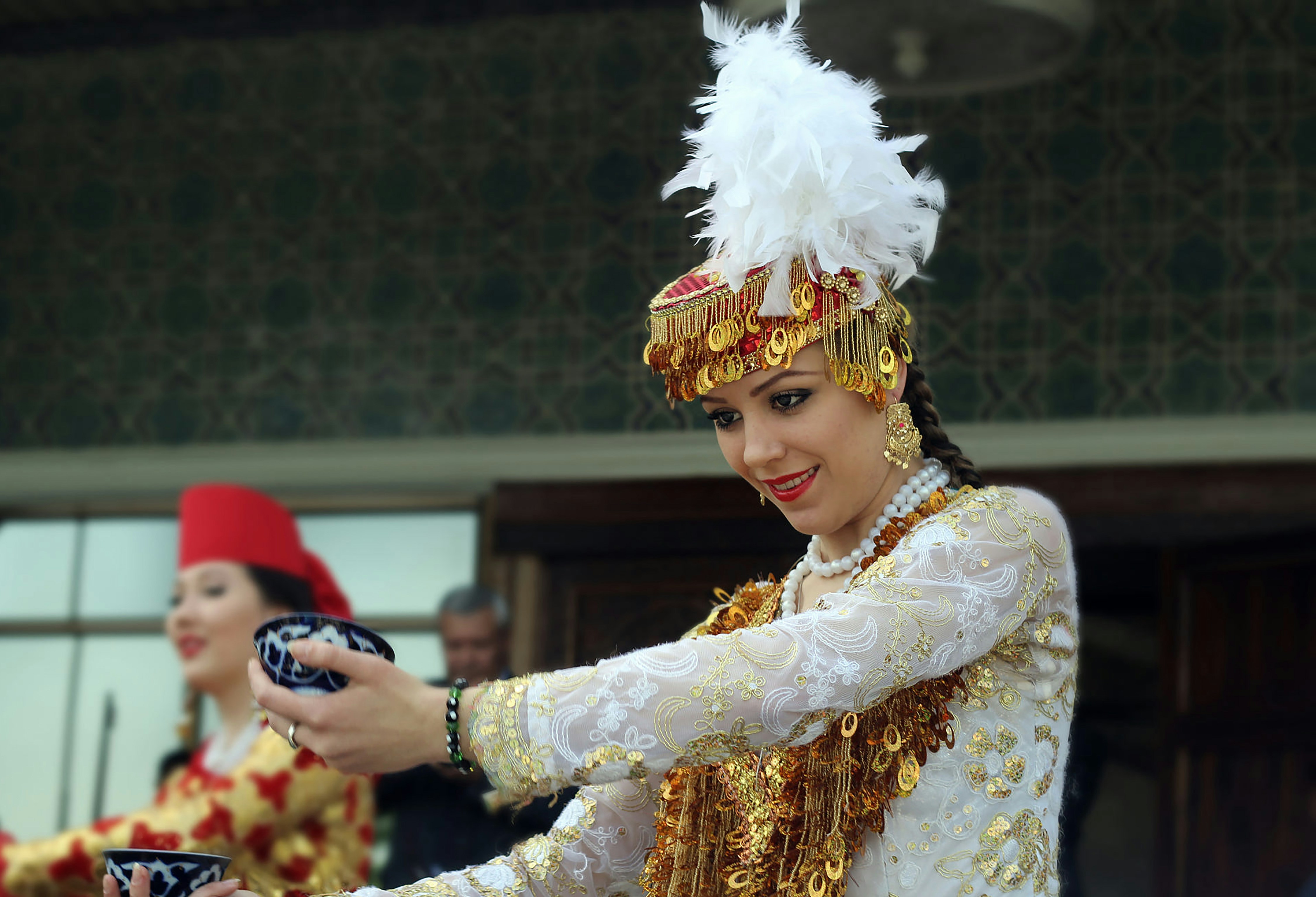 A woman in embroidered traditional dress holds a patterned bowl during a tea service.